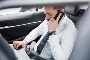 un giovane uomo d'affari che lavora al computer portatile e parla al telefono mentre è seduto nella parte posteriore dell'auto. lavora in movimento, apprezza il suo tempo