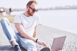 giovane seduto con il computer portatile fuori dall'ufficio. studenti che imparano online. giornata di sole estivo foto