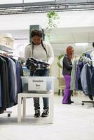 africano americano cliente la donazione Abiti per benessere organizzazione nel shopping centro. donna mettendo nuovo e secondo mano abbigliamento nel donazione scatola mentre shopping nel di moda memorizzare foto