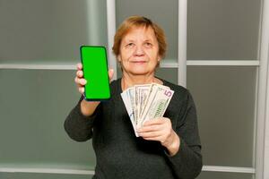 anziano donna Tenere banconote nel sua mani e mostrando un' bancario applicazione su sua Telefono foto