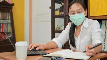la donna asiatica sta prendendo appunti e sta usando un laptop. lavora a casa per proteggersi dal virus corona o covid-19.