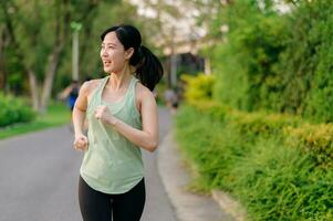 in forma asiatico giovane donna jogging nel parco sorridente contento in esecuzione e godendo un' salutare all'aperto stile di vita. femmina pareggiatore. fitness corridore ragazza nel pubblico parco. salutare stile di vita e benessere essere concetto foto