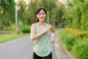 in forma asiatico giovane donna jogging nel parco sorridente contento in esecuzione e godendo un' salutare all'aperto stile di vita. femmina pareggiatore. fitness corridore ragazza nel pubblico parco. salutare stile di vita e benessere essere concetto foto