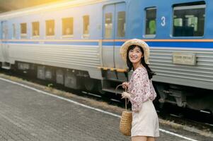 giovane asiatico donna viaggiatore con tessitura cestino guardare indietro contento sorridente per un' telecamera accanto treno In arrivo sfondo. viaggio viaggio stile di vita, mondo viaggio esploratore o Asia estate turismo concetto. foto