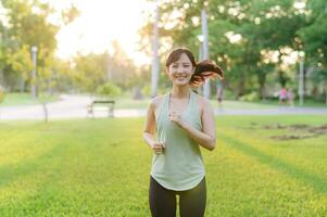in forma asiatico giovane donna jogging nel parco sorridente contento in esecuzione e godendo un' salutare all'aperto stile di vita. femmina pareggiatore. fitness corridore ragazza nel pubblico parco. salutare stile di vita e benessere essere concetto foto