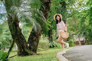 ritratto di asiatico giovane donna viaggiatore con tessitura cappello e cestino contento Sorridi su verde pubblico parco natura sfondo. viaggio viaggio stile di vita, mondo viaggio esploratore o Asia estate turismo concetto. foto