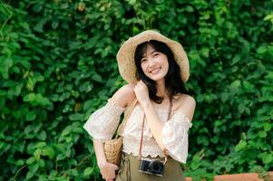 ritratto di asiatico giovane donna viaggiatore con tessitura cappello e cestino e un' telecamera su verde pubblico parco natura sfondo. viaggio viaggio stile di vita, mondo viaggio esploratore o Asia estate turismo concetto. foto