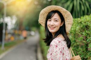 ritratto di asiatico giovane donna viaggiatore con tessitura cappello e cestino contento Sorridi su verde pubblico parco natura sfondo. viaggio viaggio stile di vita, mondo viaggio esploratore o Asia estate turismo concetto. foto