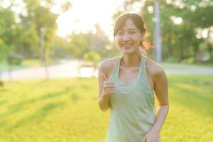 femmina pareggiatore. in forma giovane asiatico donna con verde abbigliamento sportivo allungamento muscolo nel parco prima in esecuzione e godendo un' salutare all'aperto. fitness corridore ragazza nel pubblico parco. benessere essere concetto foto