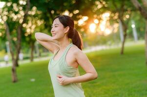 femmina pareggiatore. in forma giovane asiatico donna con verde abbigliamento sportivo allungamento muscolo nel parco prima in esecuzione e godendo un' salutare all'aperto. fitness corridore ragazza nel pubblico parco. benessere essere concetto foto