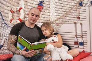 bello tatuato uomo è la spesa tempo con il suo poco carino figlia. lettura fiabe mentre figlia è seduta qui vicino. contento famiglia. foto