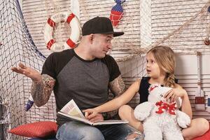 bello tatuato uomo è la spesa tempo con il suo poco carino figlia. lettura fiabe mentre figlia è seduta qui vicino. contento famiglia. foto