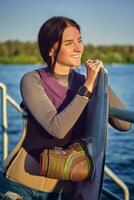 ragazza nel un' grigio dolcevita, speciale gli sport veste e nero orologio è guardare lontano e sorridente mentre in posa con sua wakeboard su un' molo. avvicinamento. foto