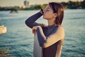 brunetta ragazza nel un' grigio dolcevita ha chiuso sua occhi mentre in posa con sua wakeboard e in piedi su un' molo di il lungo il fiume. avvicinamento. foto