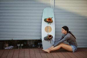 brunetta ragazza nel grigio dolcevita e denim pantaloncini è in posa con sua wakeboard seduta su pavimento a un' costiero zona. sport e ricreazione. foto