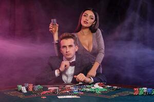 uomo giocando poker a casinò seduta a tavolo con pile di patatine fritte, i soldi, Champagne, carte. festeggiare vincere con donna. nero sfondo. avvicinamento. foto