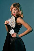 bionda ragazza nel nero elegante vestito Tenere alcuni i soldi e carte, in posa contro blu sfondo. gioco d'azzardo intrattenimento, poker, casinò. avvicinamento. foto