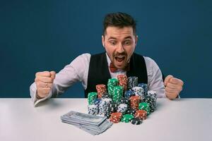 uomo nel nero veste e camicia seduta a bianca tavolo con pile di patatine fritte e denaro contante su esso, in posa su blu sfondo. gioco d'azzardo, poker, casinò. avvicinamento. foto