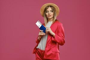 bionda modello nel cannuccia cappello, bianca camicetta e rosso tailleur pantalone. lei è Tenere passaporto e biglietto mentre in posa contro rosa studio sfondo. avvicinamento foto