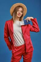 bionda modello nel cannuccia cappello, bianca camicetta e rosso tailleur pantalone. lei sorridente, mostrando passaporto e biglietto mentre in posa su blu studio sfondo. avvicinamento foto