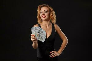 Zenzero ragazza nel buio vestito in posa Tenere fan di cento dollari fatture nel sua mani in piedi contro un' nero studio sfondo. casinò, poker. avvicinamento. foto