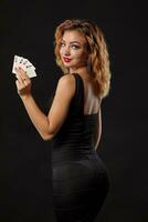 Zenzero ragazza indossare un' buio vestito è in posa Tenere quattro assi nel sua mani in piedi contro un' nero studio sfondo. casinò, poker. avvicinamento. foto