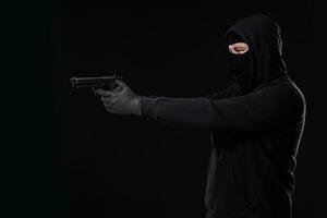 il rapinatore nel un' maschera con un' pistola appuntito per il lato su un' nero sfondo foto