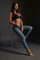 bellissimo sexy brunetta donna in posa nel studio, seduta, guardare a telecamera. ragazza indossare alla moda jeans e sensuale biancheria intima. foto