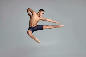 foto di un' bello uomo balletto ballerino, vestito nel un' nero corti, fabbricazione un' danza elemento contro un' grigio sfondo nel studio.