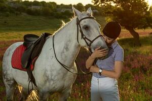 fantino giovane ragazza petting e abbracciare bianca cavallo nel sera tramonto foto