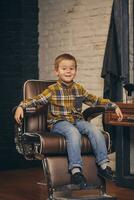 ritratto di un' elegante poco ragazzo vestito nel camicia e jeans nel il barbiere, seduta nel un' sedia contro il del barbiere posto di lavoro foto