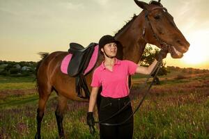 bellissimo sorridente ragazza fantino In piedi Il prossimo per sua Marrone cavallo indossare speciale uniforme su un' cielo e verde campo sfondo su un' tramonto. foto