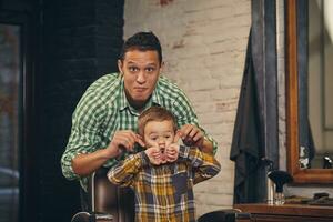 elegante poco ragazzo seduta su sedia a barbiere con il suo giovane padre su sfondo foto