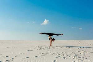 giovane donna praticante handstand su spiaggia con bianca sabbia e luminosa blu cielo foto