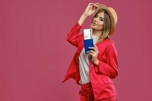 bionda signora nel cannuccia cappello, bianca camicetta e rosso tailleur pantalone. sorridente, Tenere passaporto e biglietto mentre in posa contro rosa studio sfondo. avvicinamento foto