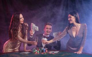 uomo giocando poker a casinò seduta a tavolo con pile di patatine fritte, i soldi, carte. festeggiare vincere con Due donne. Nero, Fumo sfondo. avvicinamento. foto