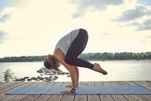 donna nel bianca maglietta e nero ghette è praticante yoga l'esecuzione yoga-asana su un' stuoia all'aperto, vicino un' lungo il fiume. foto