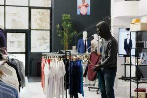 africano americano ladro indossare cappuccio, provando per spione rubato di moda merce a partire dal moderno boutique. rapinatore indossare occhiali da sole mentre furto alla moda Abiti nel shopping centro. crimine concetto foto