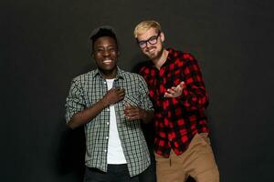 studio tiro di Due elegante giovane uomini avendo divertimento. bello barbuto fricchettone nel un' camicia nel un' gabbia in piedi Il prossimo per il suo afroamericano amico nel cappello contro un' buio sfondo. foto