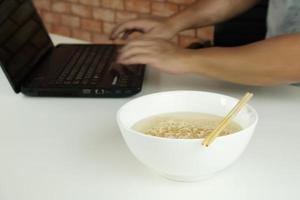 lavoratore di sesso maschile tailandese impegnato a lavorare con il laptop, usa le bacchette per mangiare frettolosamente noodles istantanei durante la pausa pranzo in ufficio, perché veloce, gustoso ed economico. nel tempo fast food asiatico, stile di vita malsano. foto