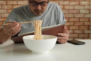 lavoratore maschio asiatico legge un libro degli appuntamenti mentre mangia noodles istantanei in una ciotola bianca con le bacchette sul tavolo in un ufficio di sfondo muro di mattoni durante una pausa pranzo, uno stile di vita frettolosamente malsano. foto