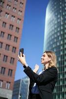 ritratto di sorridente aziendale donna video chiamata su strada, Tenere mobile Telefono e agitando a smartphone telecamera, indossare completo da uomo foto