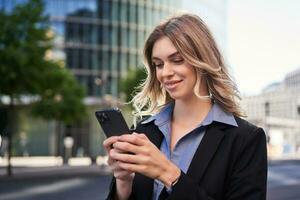 vicino su ritratto di giovane aziendale donna nel nero completo da uomo, detiene smartphone, sms Messaggio mentre in piedi su strada, utilizzando mobile applicazione foto