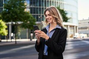 vicino su ritratto di giovane aziendale donna nel nero completo da uomo, detiene smartphone, sms Messaggio mentre in piedi su strada, utilizzando mobile applicazione foto