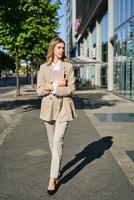 verticale tiro di donna d'affari a piedi su strada con digitale tavoletta, andando per opera, indossare beige completo da uomo e alto tacchi foto