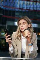 verticale tiro di donna d'affari bevande caffè, sembra a mobile Telefono app. aziendale donna su sua pranzo rompere, utilizzando smartphone foto