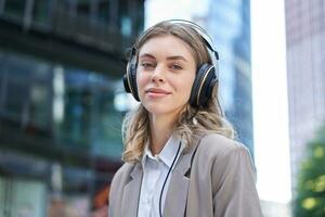 ritratto di sorridente aziendale donna, ragazza nel completo da uomo ascoltando musica nel cuffia, seduta nel città centro foto