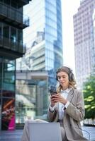 aziendale donna nel completo da uomo, seduta nel città, ascoltando per musica con computer portatile, scorrimento notizia su sua mobile Telefono, rilassante su pranzo rompere foto