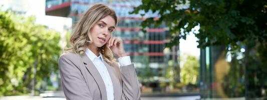 ritratto di fiducioso aziendale donna, giovane femmina Amministratore delegato manager Guarda a telecamera, in posa nel beige completo da uomo vicino ufficio edifici, in piedi su strada foto