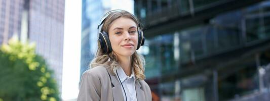 ritratto di sorridente aziendale donna, ragazza nel completo da uomo ascoltando musica nel cuffia, seduta nel città centro foto